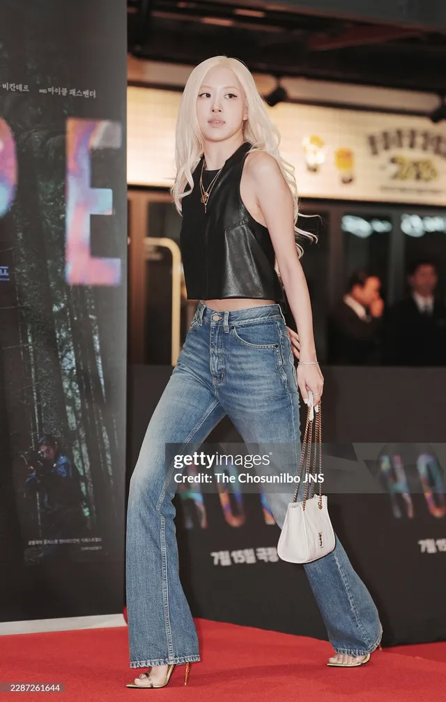 Rosé standing at a premiere in a black top and blue jeans while carrying a small handbag.