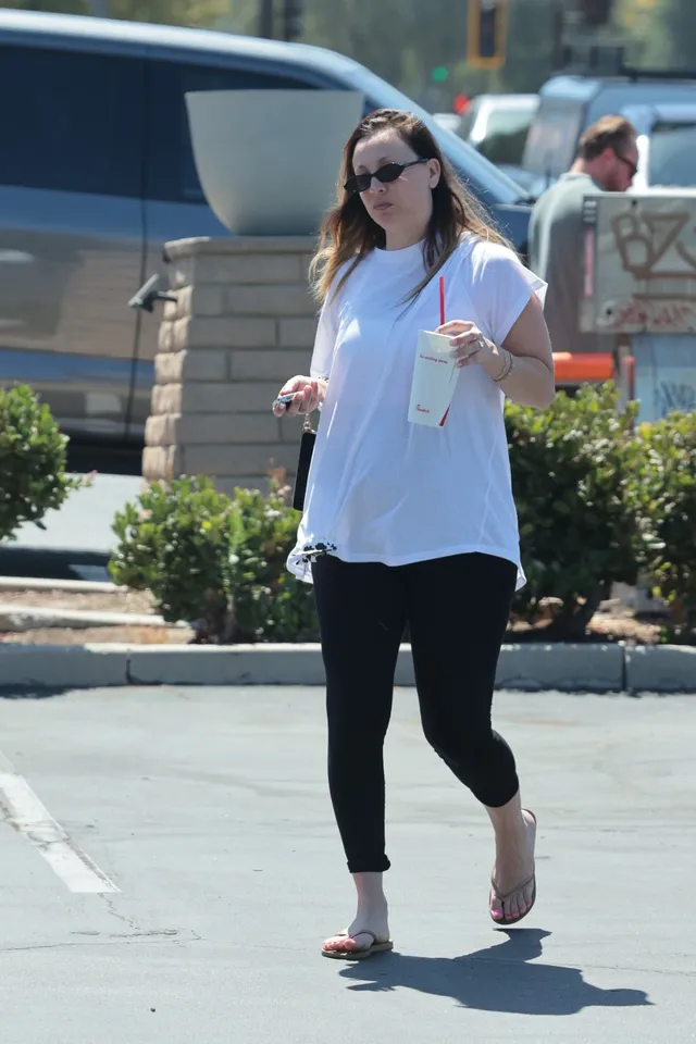 Kaley Cuoco standing outdoors in a white blouse and flip-flops.