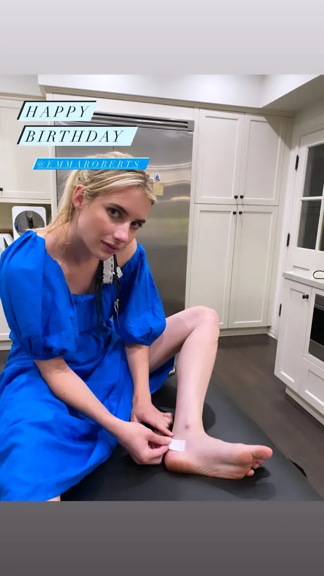 Emma Roberts sitting barefoot on a dark counter in a blue dress.