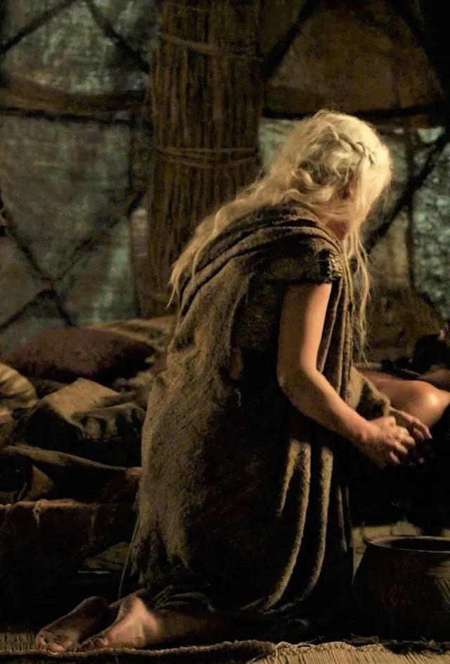 Emilia Clarke kneeling barefoot beside a fire in a film scene.