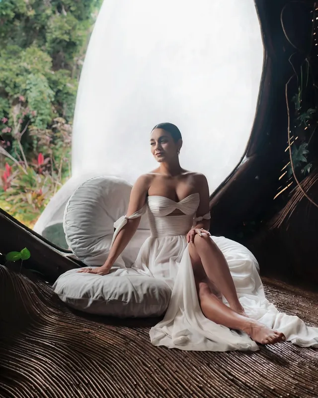 Vanessa Hudgens sitting barefoot in a chair wearing a pale gown.