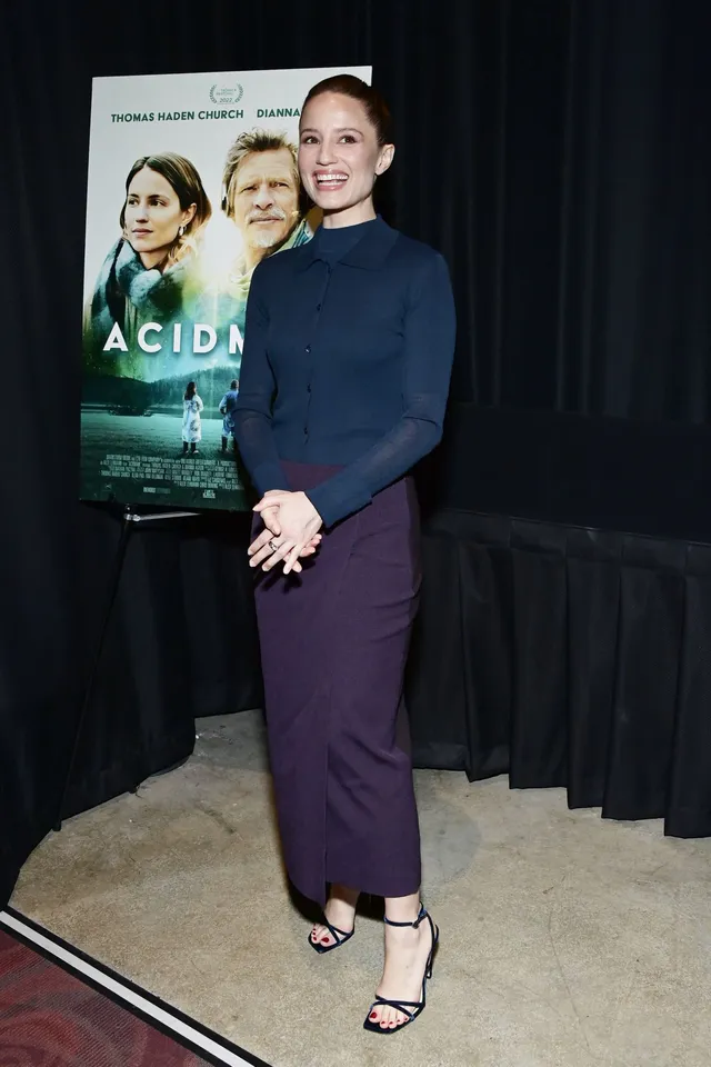 Dianna Agron standing beside a film poster in a dark outfit.