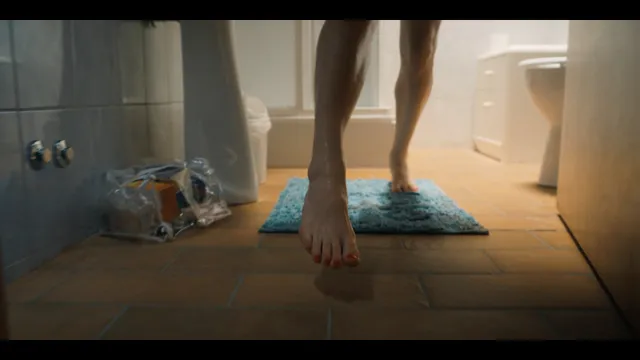 Grace Van Patten walking barefoot across a tiled bathroom floor in a film scene.