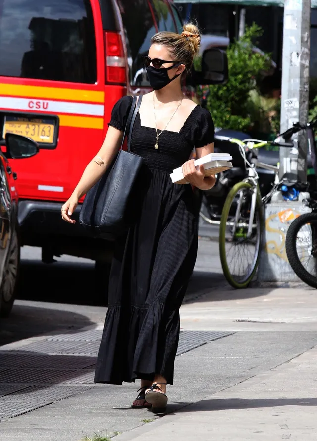 Dianna Agron walking on a pavement in a black dress and sandals.