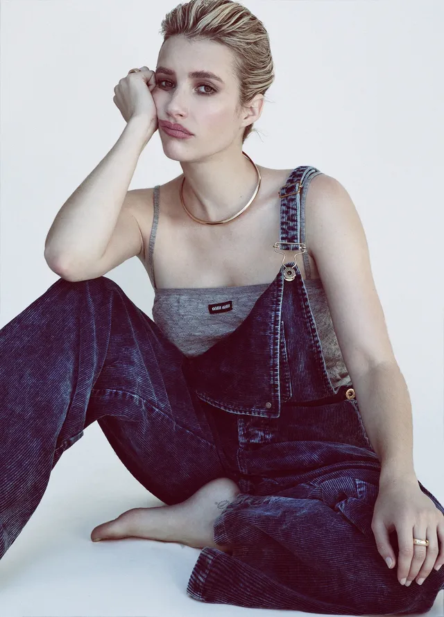 Emma Roberts sitting barefoot in denim overalls for a portrait.