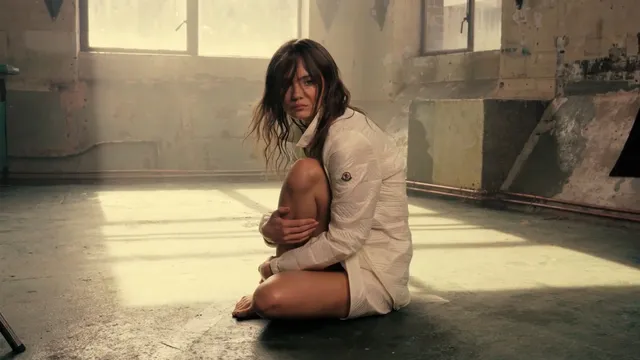 Freya Allan sitting barefoot in a pale outfit on a studio floor.