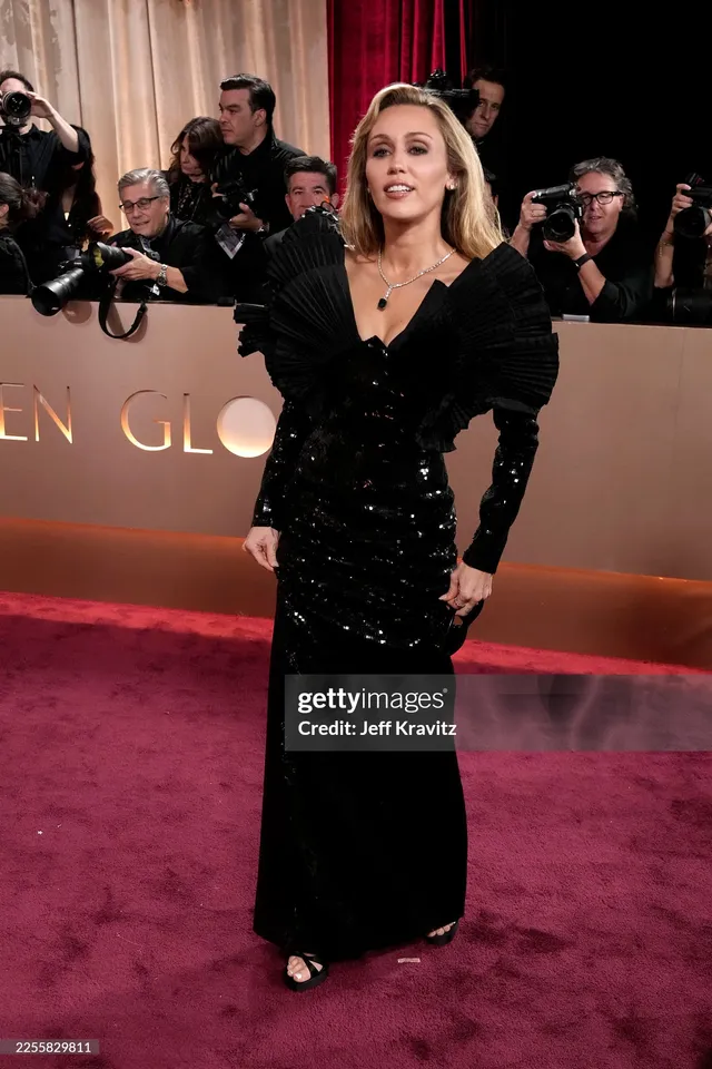 Miley Cyrus posing on a carpet in a black dress and heels.