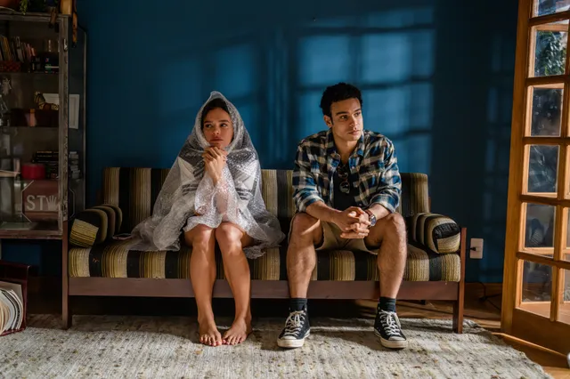 Bruna Marquezine sitting barefoot beside a man on a sofa in a scene.