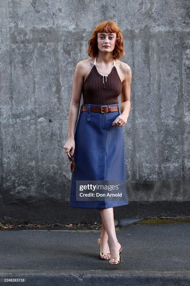 Ella Purnell posing against a grey wall in a blue skirt.