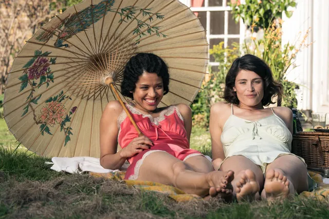 Gemma Arterton sitting outdoors beside another woman and a parasol.