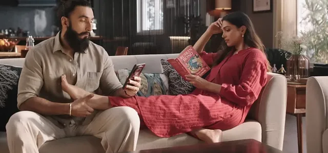 Deepika Padukone showing flat sandals while reclining beside a companion on a sofa.