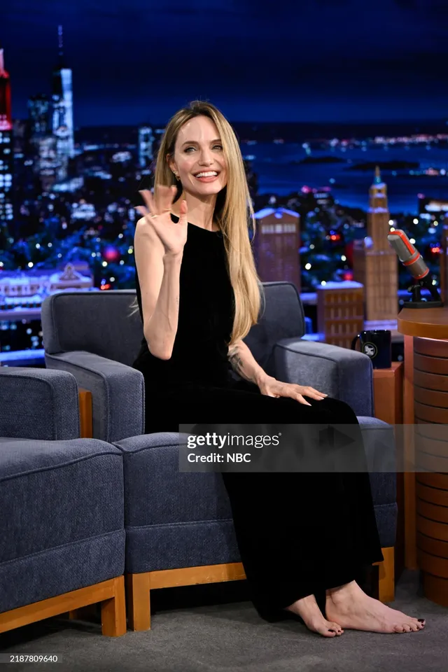 Angelina Jolie seated on a talk show chair in a long black dress.
