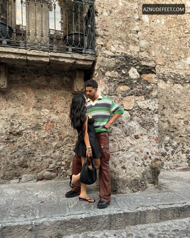 Cierra Ramirez in a dark casual outfit and flat sandals with her partner beside a stone wall.
