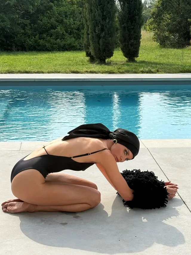 Elsa Hosk kneeling barefoot beside a pool in a black swimsuit.