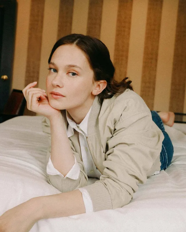 Cailee Spaeny lying barefoot on a bed in a pale jacket.