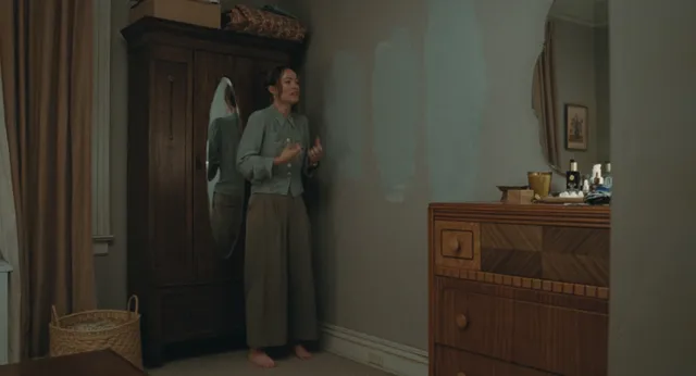 Olivia Wilde standing barefoot beside a dresser in a film scene.