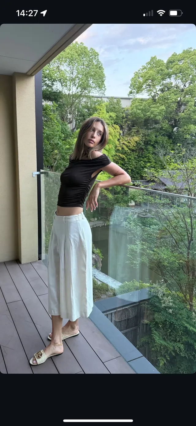 María Pedraza posing outdoors in a dark top, white skirt and flat sandals.