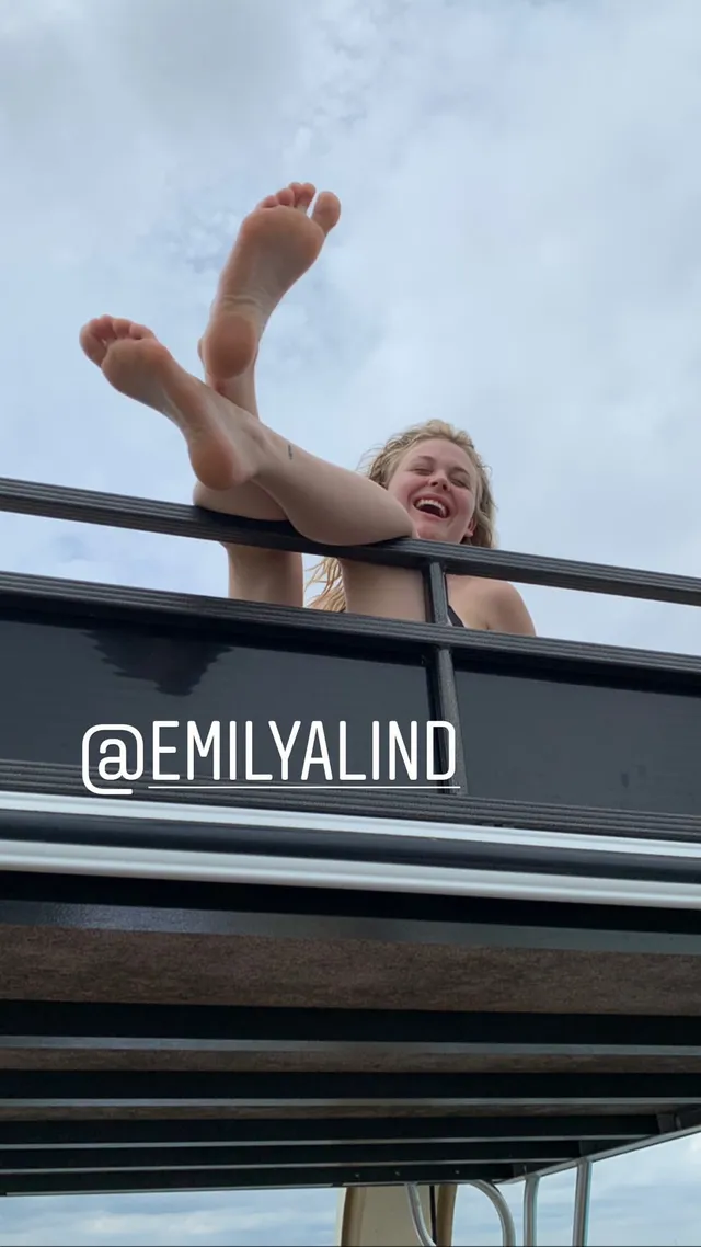 Emily Alyn Lind leaning over a railing with bare feet raised outdoors.