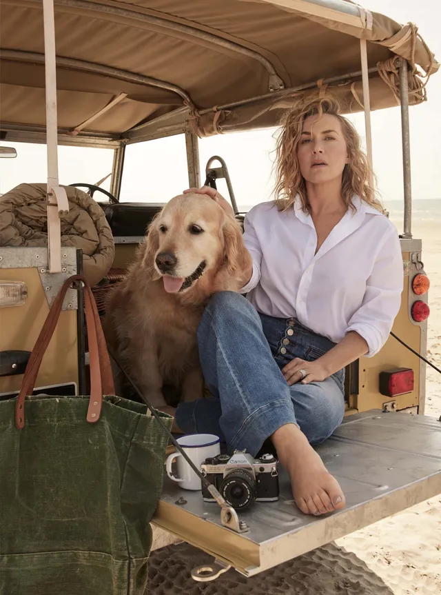 Kate Winslet sitting barefoot on a vehicle beside a dog.