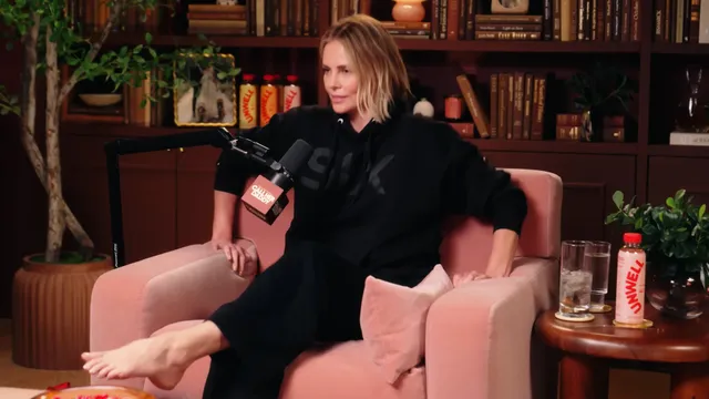 Charlize Theron seated on a talk show chair in a black outfit.
