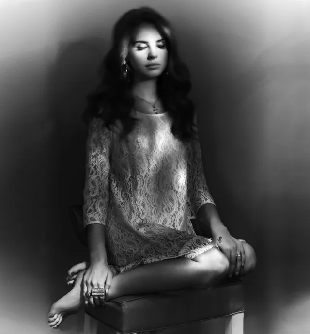 Lana Del Rey sitting cross-legged on a stool in a monochrome portrait.