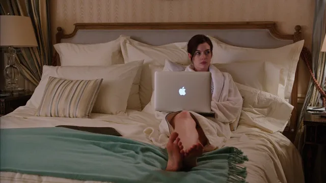 Anne Hathaway sitting on a bed with bare soles visible beside a laptop.