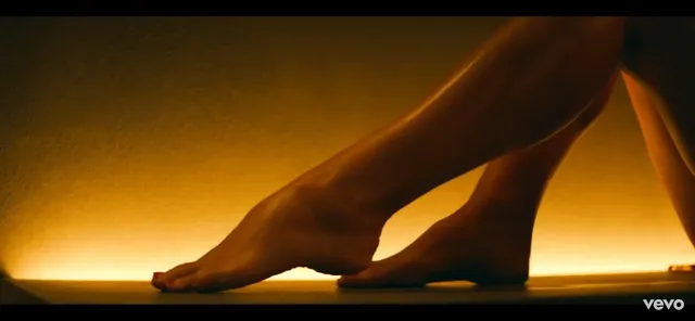 Lana Del Rey standing barefoot in a warmly lit video scene.