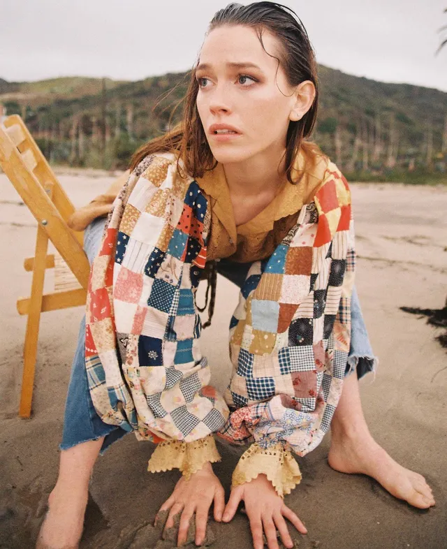 Victoria Pedretti sitting on a beach in a patchwork jacket.