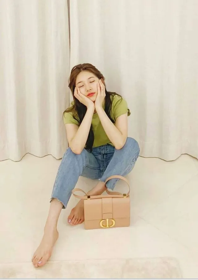 Bae Suzy sitting barefoot in jeans beside a handbag.