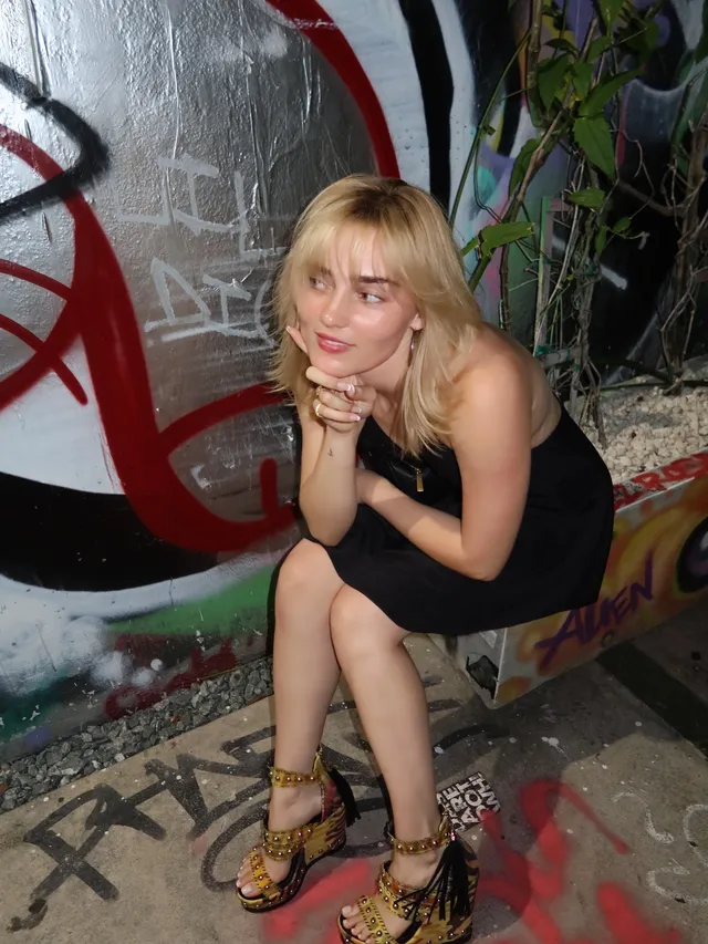 Meg Donnelly sitting beside a graffiti wall in a black dress and patterned platform heels.