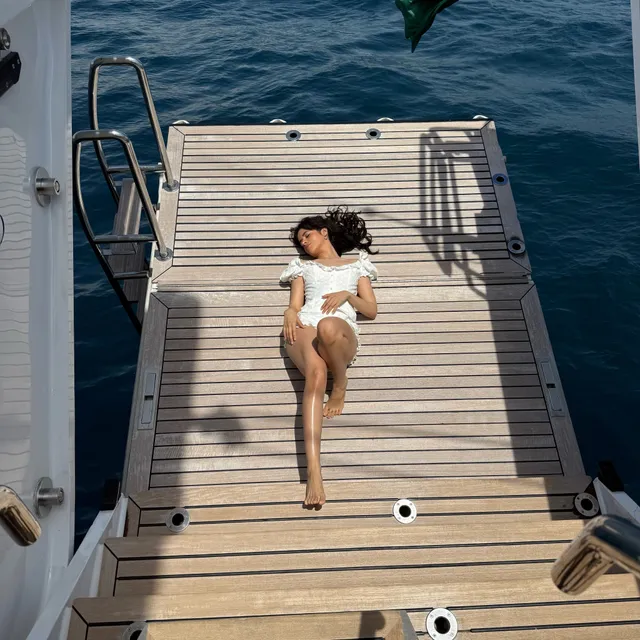 Camila Cabello lying barefoot on a boat deck in a white outfit.