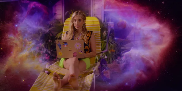 Emma Roberts seated barefoot with a laptop against a colorful backdrop.