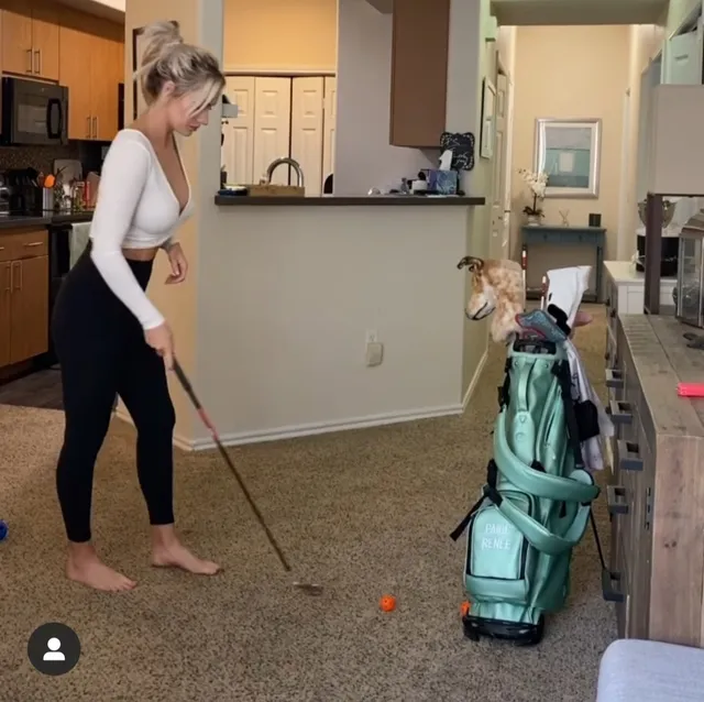 Paige Spiranac standing barefoot beside a golf bag while practising indoors.
