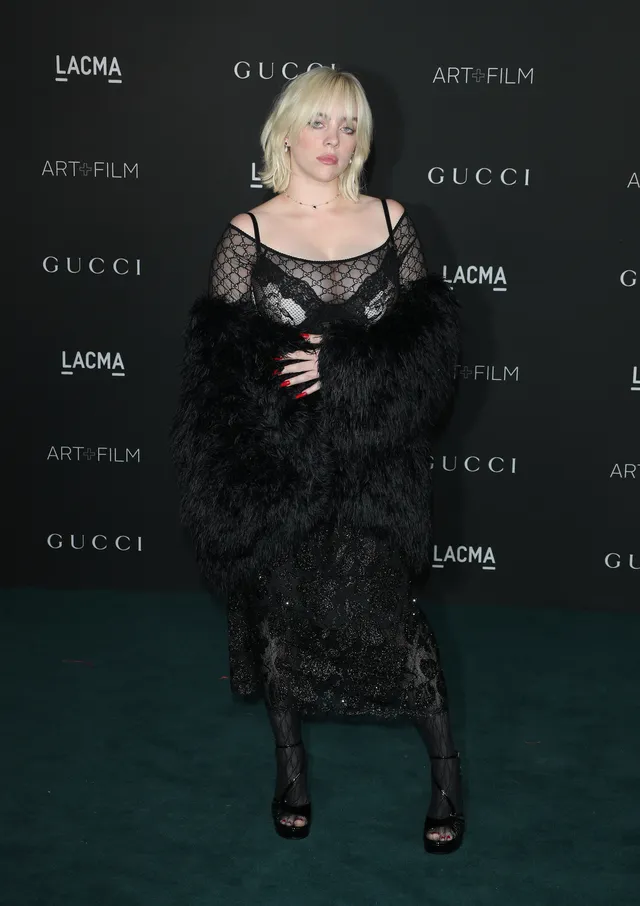 Billie Eilish posing at an event in a lace dress and dark heels.
