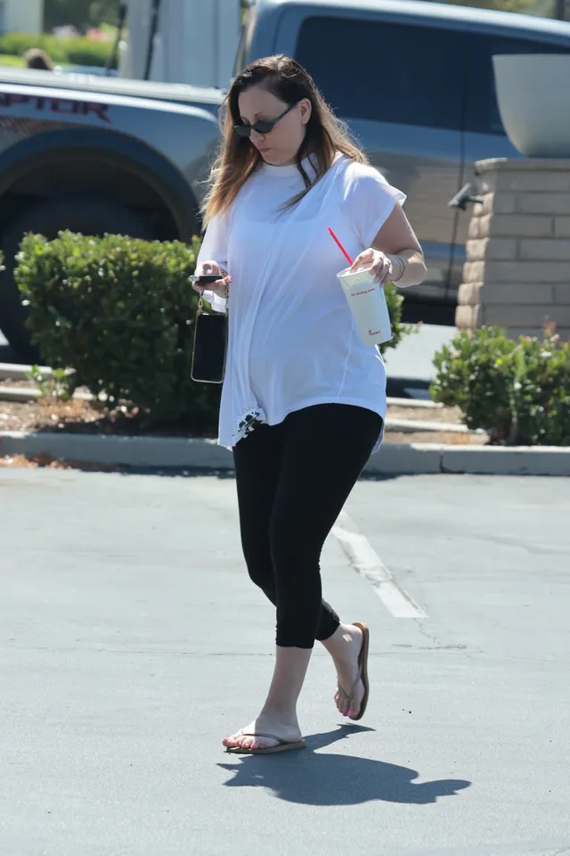 Kaley Cuoco walking outdoors in a white blouse and flip-flops.