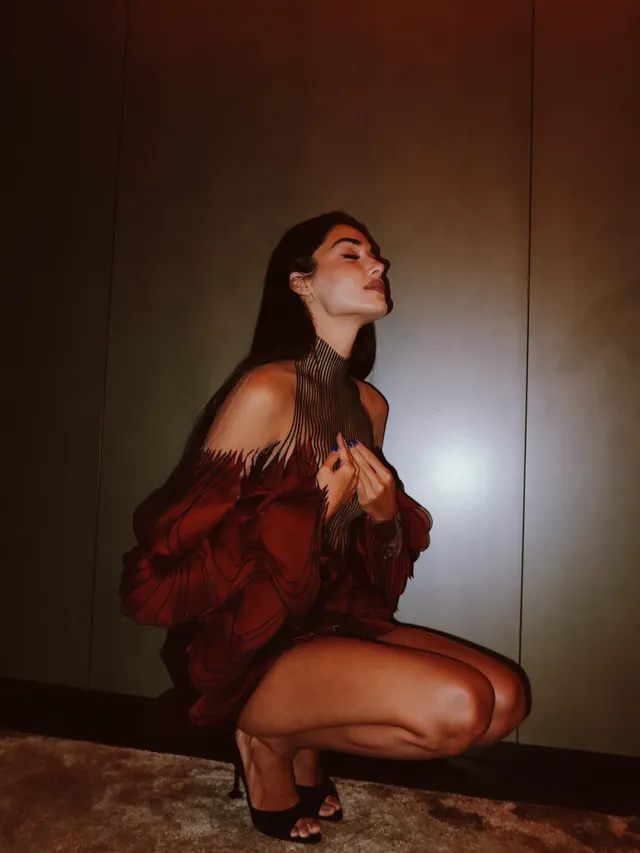 Hande Erçel crouching indoors in a rust-colored outfit and black heels.
