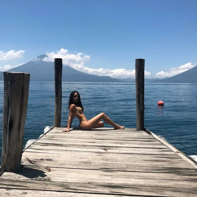 Adria Arjona posing barefoot on a jetty with mountains behind her.