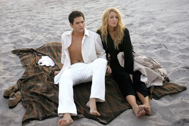 Blake Lively sitting barefoot beside a companion on beach sand.