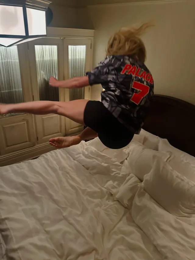 Ellie Goulding jumping barefoot on a bed in a dark shirt and shorts.