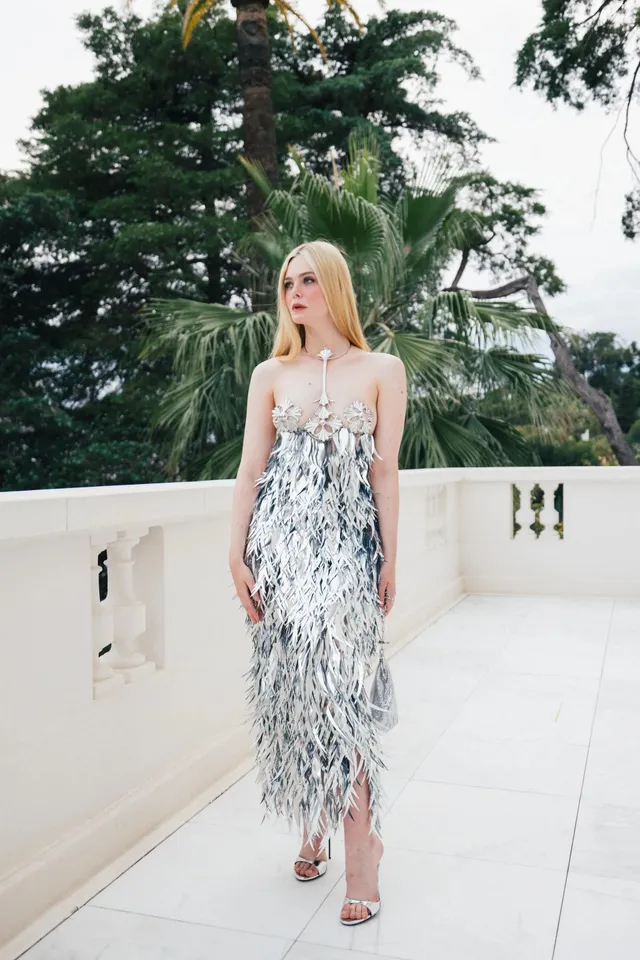 Elle Fanning standing on a terrace in a silver dress and heels.