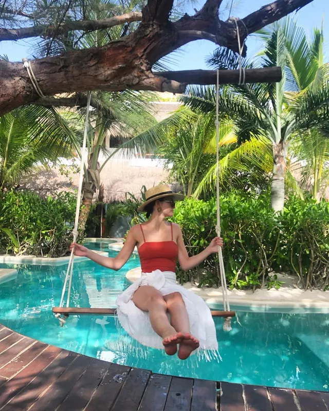 Maia Mitchell sitting barefoot on a swing over a swimming pool.