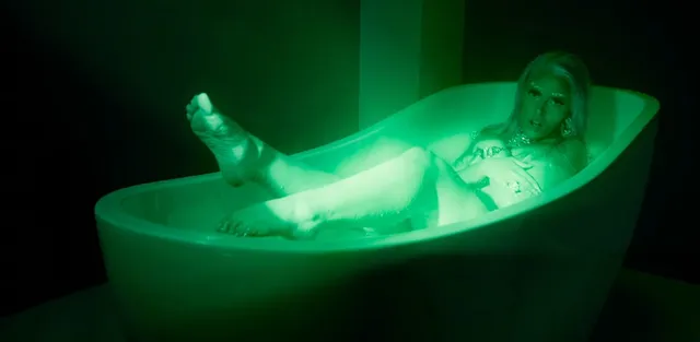 Doja Cat reclining in a bathtub under green lighting with bare feet raised.