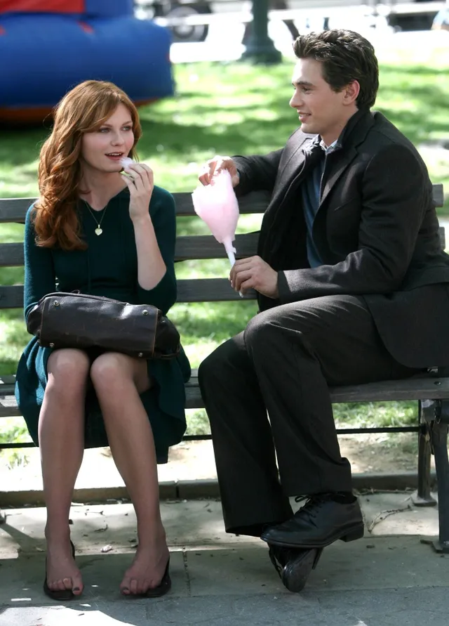 Kirsten Dunst sitting barefoot on a bench beside another person.