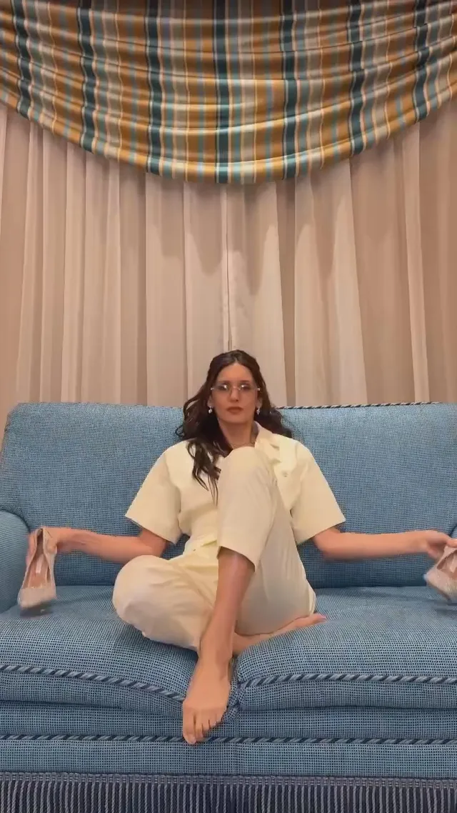 Nina Dobrev sitting barefoot on a blue sofa in a pale outfit.