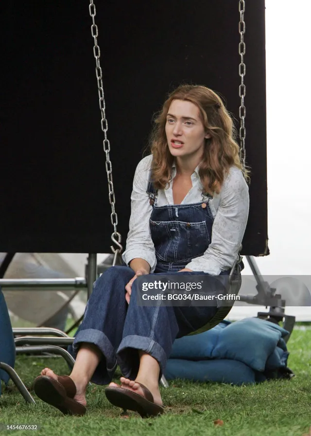 Kate Winslet sitting on grass in jeans and sandals.