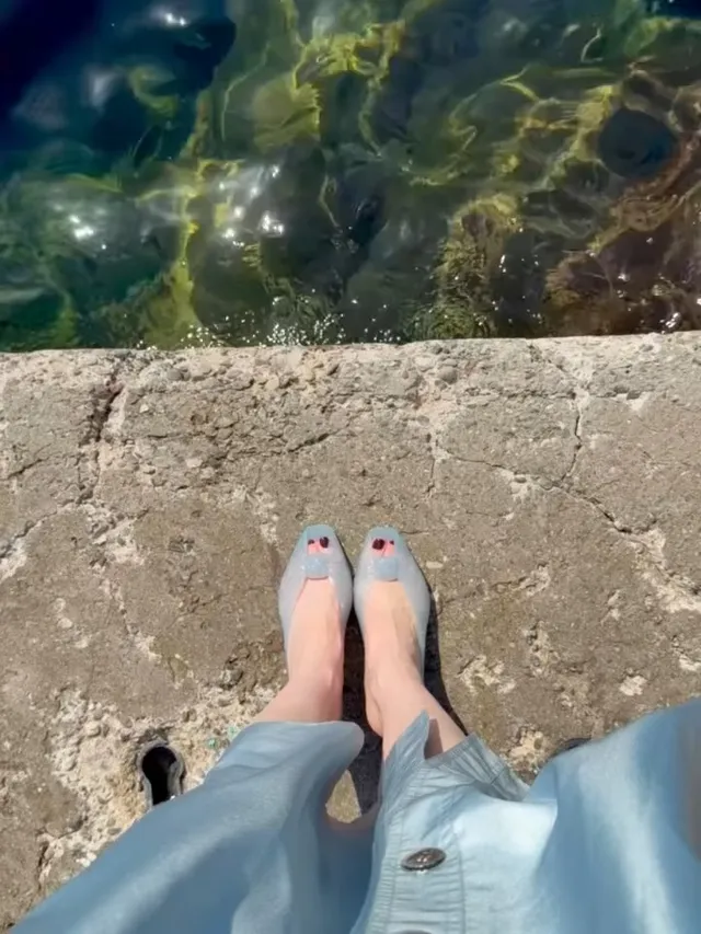 Lily Collins standing beside clear water wearing translucent flats.