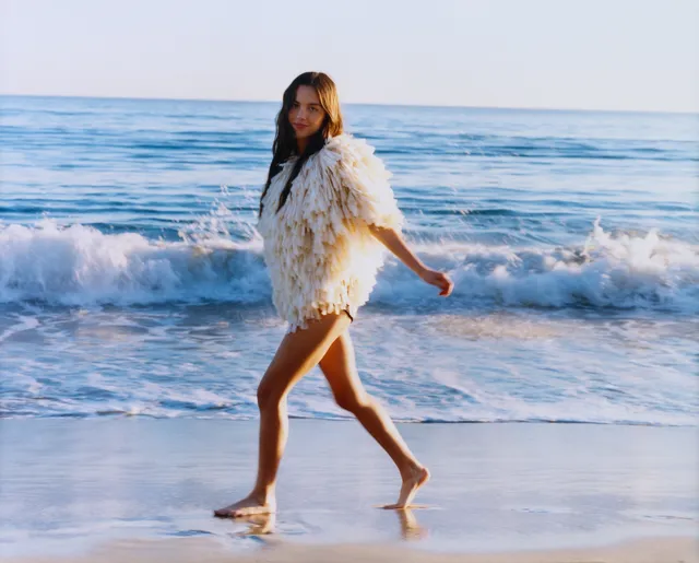 Olivia Rodrigo walking barefoot along the shoreline in a pale jacket.