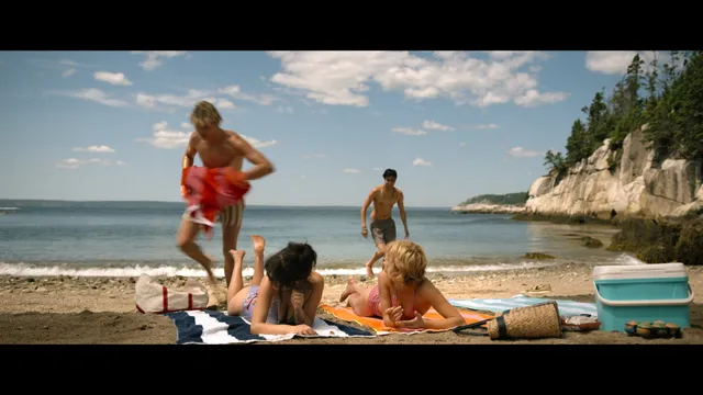 Emily Alyn Lind sitting barefoot with a group beside the sea.