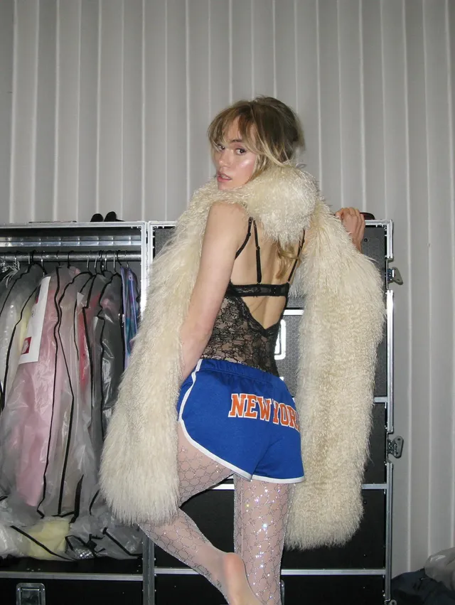 Suki Waterhouse posing in a dressing room in a long pale coat.