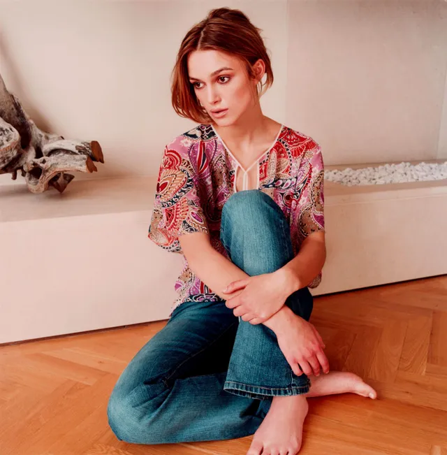 Keira Knightley seated on a wooden floor in jeans and a patterned top.